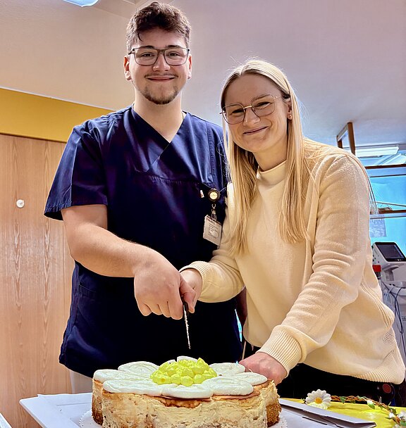 Zwei lächelnde Personen schneiden gemeinsam eine große, runde Torte an. Der links Stehende trägt dunkelblaue Arbeitskleidung, die rechts Stehende einen hellen Pullover. Auf der Torte liegen helle Obststücke. Fokus: gemeinsamer Anschnitt bei einer Feier.