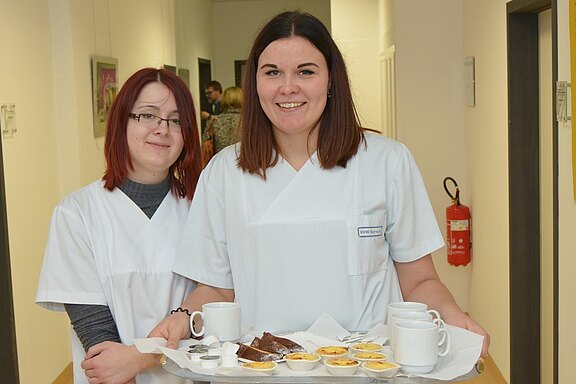 Zwei Personen in weißen Pflegeuniformen stehen in einem Flur. Die eine Person hält ein Tablett mit Tassen und kleinen Desserts. Der Hintergrund zeigt eine einfache Wand und einige Personen in der Ferne.