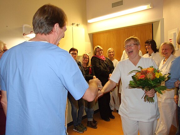 In einem Krankenhausflur gratuliert ein Mitarbeiter einer lachenden Pflegekraft per Handschlag; sie hält einen Blumenstrauß. Kolleginnen und Kollegen lächeln im Hintergrund.