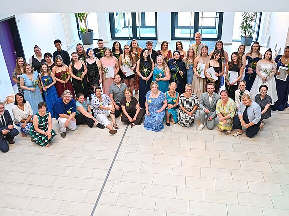 Großes Gruppenfoto in einem hellen Innenraum: Erwachsene in festlicher Kleidung stehen und sitzen zusammen, lächeln zur Kamera. Viele halten Urkunden und Blumen. Die Szene wirkt wie eine Abschluss- oder Ehrungsfeier.