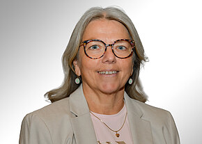 Margit Krieger Porträt einer lächelnden Frau mittleren bis höheren Alters mit grauen, schulterlangen Haaren und großer Brille. Sie trägt einen beigefarbenen Blazer, rosafarbenes Oberteil und dezente Ohrringe vor neutralem Hintergrund.