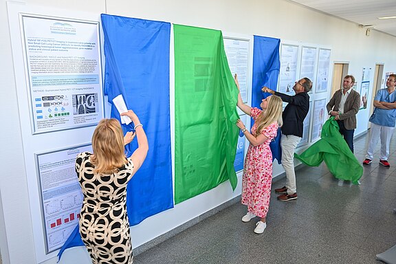 Die Abbildung zeigt eine Gruppe von Personen, die farbige Stoffe (blau und grün) von Informationsständen abziehen. Diese Stände enthalten Forschungsprojekte zur Hybrid-PET-CT-Bildgebung bei nicht kleinzelligem Lungenkrebs. Die Anwesenden wirken interessiert und engagiert.