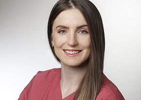 Porträt einer lächelnden Frau mit langen, glatten Haaren in einem rosa Oberteil. Der Hintergrund ist hell und neutral. Die Frau hat grüne Augen und trägt kleine Ohrringe. Sie strahlt Freundlichkeit und Professionalität aus.