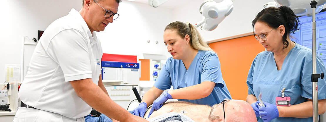 Drei medizinische Fachkräfte führen eine Untersuchung an einem Patienten durch, der auf einer Liege liegt. Der Patient ist teilweise entblößt, und einer der Helfer befestigt Elektroden an seiner Brust. Die Umgebung wirkt modern und klinisch.