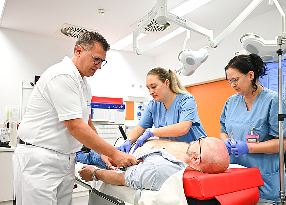 Drei medizinische Fachkräfte führen eine Untersuchung an einem Patienten durch, der auf einer Liege liegt. Der Patient ist teilweise entblößt, und einer der Helfer befestigt Elektroden an seiner Brust. Die Umgebung wirkt modern und klinisch.