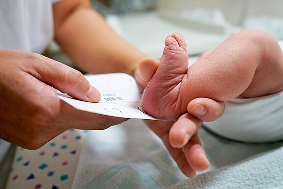 Nahaufnahme: Eine medizinische Fachkraft hält den Fuß eines Neugeborenen und führt den Fersenbluttest für das Neugeborenen-Screening mit einer Karte durch.