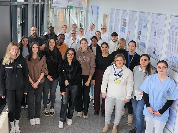 Gruppe von rund 25 Personen steht in einem hellen Flur und blickt in die Kamera. Einige tragen medizinische Kleidung oder Laborkittel, andere Freizeitkleidung. An den Wänden hängen Poster, die auf eine klinische oder wissenschaftliche Einrichtung hinweisen.