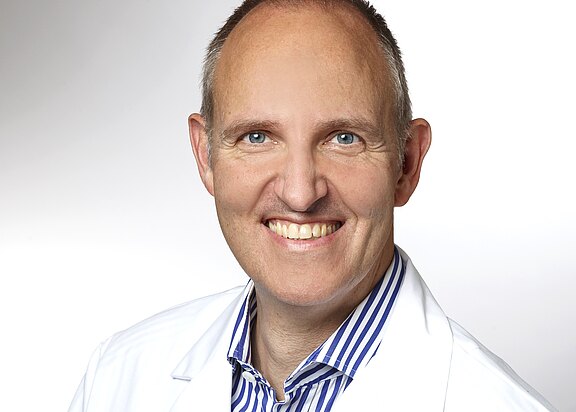 Porträt eines lächelnden Mannes im weißen Arztkittel vor hellem Hintergrund; vermittelt Professionalität und Freundlichkeit.