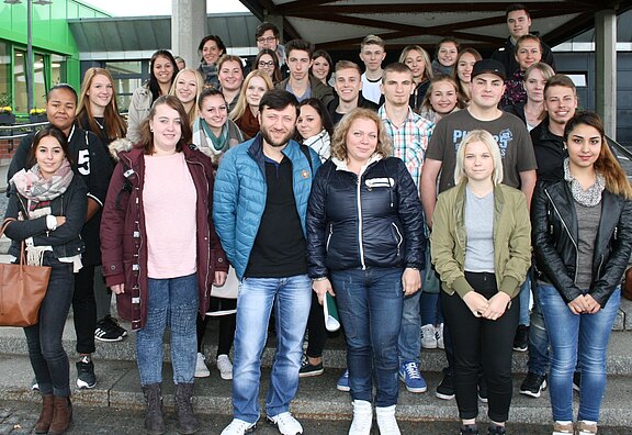 Gruppe von rund 30 Jugendlichen und wenigen Erwachsenen posiert auf Treppenstufen für ein Gruppenfoto vor einem Gebäude; alle blicken in die Kamera.