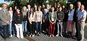 Gruppenfoto mit einer Vielzahl von Personen, sowohl Männern als auch Frauen, die sich im Freien in einer parkähnlichen Umgebung versammeln. Die Anwesenden lächeln und stehen in lockerer Aufstellung, während einige Bäume im Hintergrund sichtbar sind.