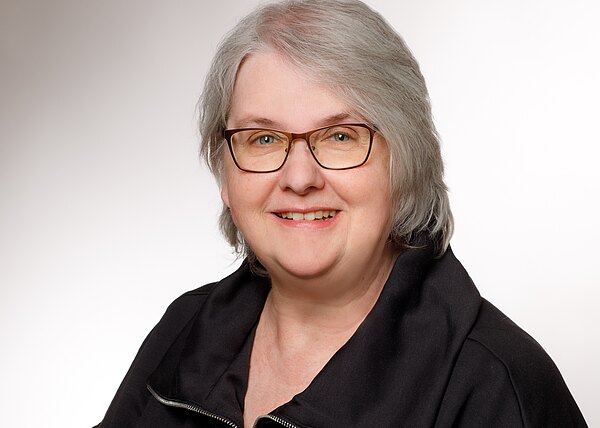 Porträt einer lächelnden erwachsenen Person mit schulterlangem grauem Haar und Brille, in dunkler Jacke, vor neutralem Hintergrund; direkter Blick in die Kamera.