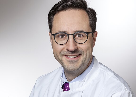 Porträt eines mittelalten Mannes mit Brille im weißen Arztkittel. Er lächelt freundlich in die Kamera; violette Krawatte sichtbar. Neutraler, heller Hintergrund.