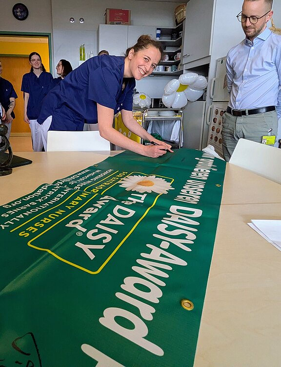 Eine Pflegekraft in blauer Dienstkleidung unterschreibt lächelnd ein großes grünes Banner mit der Aufschrift „DAISY Award“ und einer Gänseblume. Kolleginnen und Kollegen stehen daneben und schauen zu. Szene wirkt wie eine kurze Feier oder Anerkennung im Pausenraum eines Krankenhauses.