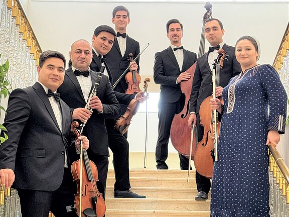 Eine Gruppe von sieben Musikern steht auf einer Treppe. Sie tragen formelle Kleidung und halten verschiedene Musikinstrumente, darunter Geigen, ein Cello und eine Flöte. Im Hintergrund ist eine elegante Treppe und ein Lichtstrahl zu sehen. Die Musiker wirken professionell und sind bereit zu spielen.