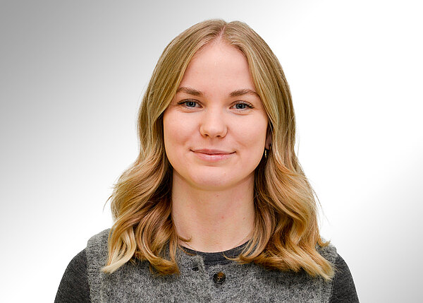 Porträtfoto: Junge Frau mit schulterlangen, leicht welligen blonden Haaren blickt in die Kamera und lächelt dezent. Sie trägt ein graues Oberteil. Heller, neutraler Hintergrund.