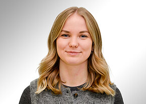 Porträtfoto: Junge Frau mit schulterlangen, leicht welligen blonden Haaren blickt in die Kamera und lächelt dezent. Sie trägt ein graues Oberteil. Heller, neutraler Hintergrund.