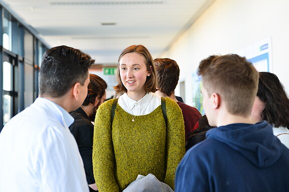 Mehrere junge Erwachsene unterhalten sich in einem hellen Flur. Im Mittelpunkt steht eine Frau mit grünem Pullover und Rucksackriemen, die aufmerksam einem Mann gegenüber zuhört. Die Szene wirkt wie ein informelles Gespräch bei einer Veranstaltung oder in einer Bildungseinrichtung.