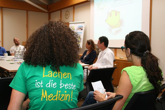 Gruppe in einem Besprechungsraum bei einer Diskussion. Im Vordergrund sitzt eine Person mit lockigen Haaren im grünen T‑Shirt mit dem Aufdruck „Lachen ist die beste Medizin!“. Im Hintergrund laufen Präsentationsfolien; die Teilnehmenden hören zu und sprechen.
