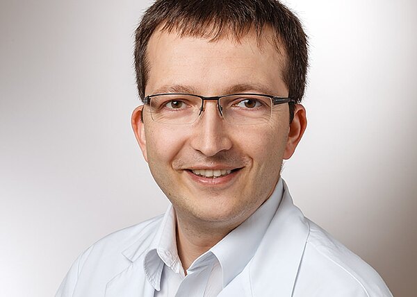 Porträt eines lächelnden Mannes mit Brille und kurzer brauner Frisur, der einen weißen Kittel trägt und in die Kamera blickt; neutraler, heller Hintergrund.