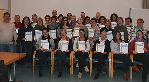 Gruppenfoto von rund 30 Erwachsenen in einem Raum. Viele sitzen vorne, andere stehen dahinter. Alle halten Zertifikate in den Händen und lächeln. Es wirkt wie der Abschluss oder die Verleihung von Teilnahmeurkunden eines Kurses oder einer Schulung.