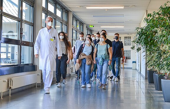 Ein Arzt im weißen Kittel führt eine Gruppe junger Erwachsener durch einen hellen Krankenhausflur. Alle tragen medizinische Masken, hören zu und gehen gemeinsam. Die Szene vermittelt Führung und Orientierung in einer Klinik.