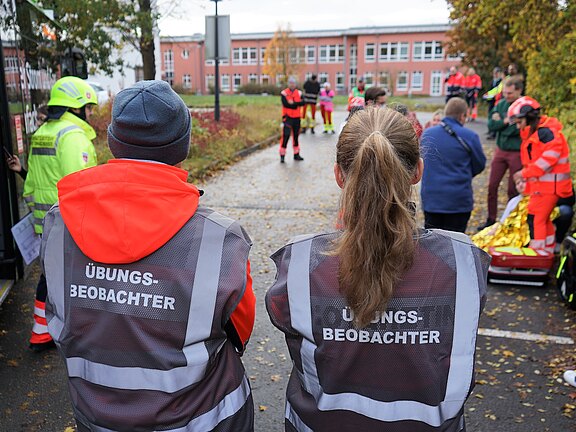 Zwei Personen in orangefarbenen Westen mit der Aufschrift "Übungs-Beobachter" stehen im Vordergrund. Im Hintergrund sind mehrere Menschen zu sehen, die an einem Einsatztraining teilnehmen. Es handelt sich um eine Übung zur Notfallversorgung.