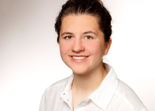 Professionelles Porträt einer freundlich lächelnden jungen Frau mit hochgesteckten Haaren in weißem Hemd vor neutral hellem Hintergrund.
