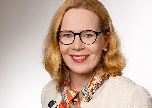 Porträtfoto einer lächelnden Frau mittleren Alters mit schulterlangem blondem Haar, runder schwarzer Brille und Perlenohrring. Sie trägt eine helle Jacke und ein buntes, überwiegend orangefarbenes Tuch. Neutraler Studiohintergrund, Kopf und Schultern im Bild.