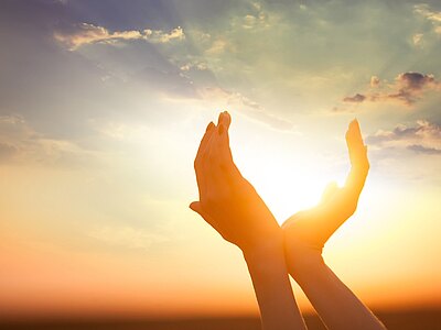 Zwei erhobene Hände formen eine Schale und halten das helle Sonnenlicht. Dahinter ein Himmel mit sanften Wolken bei Sonnenuntergang. Motiv von Wärme und Hoffnung.