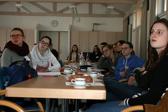 Mehrere junge Erwachsene sitzen in einem Seminarraum um Tische, mit Notizen, Kaffee und Gebäck. Sie schauen aufmerksam in eine Richtung, als hörten sie einem Vortrag oder einer Diskussion zu.