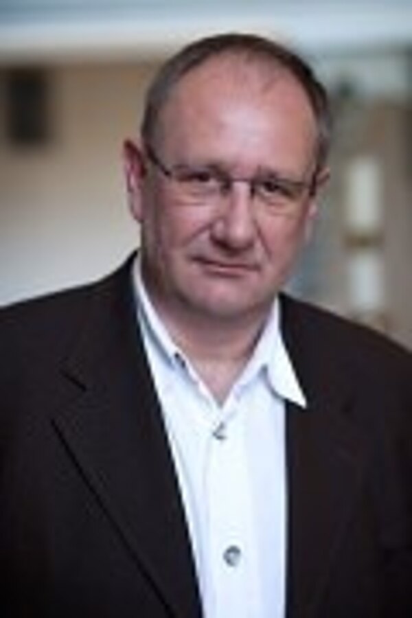 Porträt eines mittelalten Mannes mit Brille. Er trägt ein dunkles Sakko über einem weißen Hemd und blickt ernst in die Kamera. Neutraler, unscharfer Hintergrund.