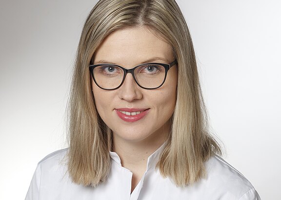 Porträt einer erwachsenen Frau mit schulterlangem blondem Haar und schwarzer Brille, freundlich lächelnd. Sie trägt einen weißen Kittel vor neutralem Hintergrund und blickt direkt in die Kamera.