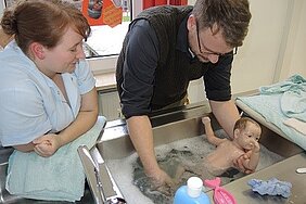 Ein Mann badet ein Säuglingsmodell in einer Spüle, während eine Frau daneben zusieht. Der Raum ist hell und mit Handtüchern sowie Reinigungsmitteln ausgestattet. Die Szene vermittelt eine Lern- oder Trainingssituation zur Pflege von Kleinkindern.