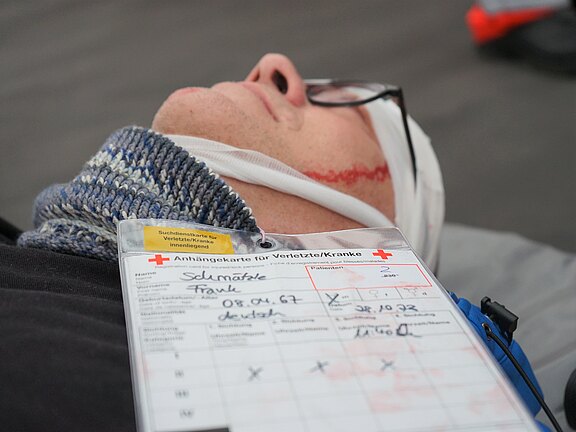 Ein verletzter Mann liegt auf dem Boden. Er trägt einen Verband um den Kopf, und auf einem Dokument neben ihm ist sein Name, Geburtsdatum und Informationen zur Verletzung vermerkt. Das Dokument gehört zu einer Krankenversorgung.