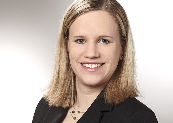 Porträtfoto einer jungen Frau mit schulterlangem blondem Haar, lächelnd, Blick zur Kamera. Sie trägt einen schwarzen Blazer und eine schlichte Halskette. Heller, neutraler Hintergrund.