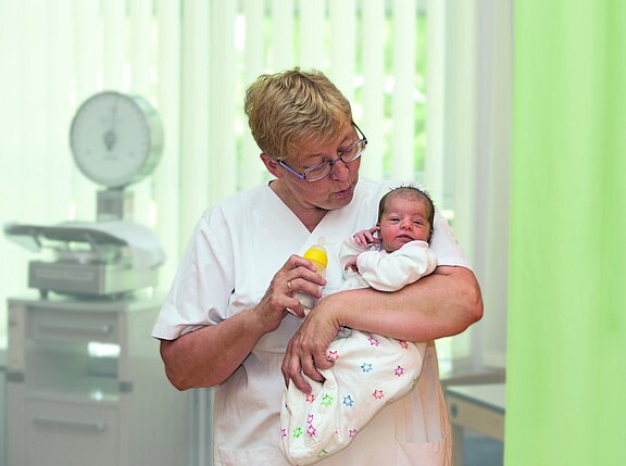 Pflegekraft in weißer Kleidung hält ein Neugeborenes im Arm und bietet eine Milchflasche an. Szene in einer Klinik, ruhig und fürsorglich.