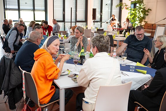 Gruppen von Erwachsenen sitzen in einem hellen Veranstaltungsraum an gedeckten Tischen, sprechen angeregt bei Getränken und Notizen – eine ungezwungene Diskussionsrunde oder ein Netzwerktreffen.