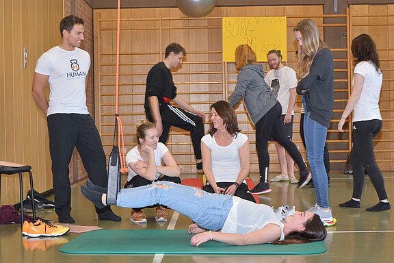 Eine Gruppe von Teilnehmern in Sportkleidung führt Übungen in einer Turnhalle durch. Eine Frau liegt auf einer Matte, während andere sie beobachten und miteinander lachen. Einige Personen sind mit Fitnessgeräten beschäftigt, und eine große Sportmatte liegt auf dem Boden.