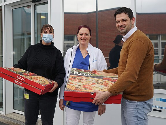 Drei Personen stehen vor einem Gebäude und halten große Pizzakartons. Eine Frau trägt eine Maske und hat ein schwarzes Oberteil an, eine zweite Frau trägt einen weißen Kittel und die dritte Person hat ein braunes Pullover. Alle lächeln und scheinen sich zu freuen.