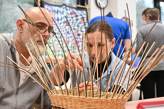 Zwei Personen flechten gemeinsam einen Korb aus Weidenruten. Sie arbeiten konzentriert, führen die Ruten zwischen die Streben und besprechen den nächsten Schritt. Werkstattatmosphäre im Hintergrund, Fokus auf Zusammenarbeit und handwerkliches Korbflechten.