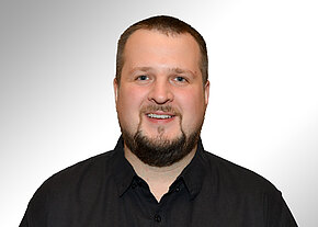 Porträt eines lächelnden erwachsenen Mannes mit kurzem Haar und Vollbart, in schwarzem Hemd vor hellem, neutralem Hintergrund, zentriert und frontal ins Bild blickend.