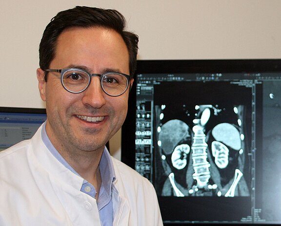 Ein Arzt im weißen Kittel lächelt neben einem Monitor mit einer medizinischen Bildgebung des Bauchraums. Auf dem Scan sind deutlich Wirbelsäule und Nieren zu sehen. Die Szene steht für Diagnostik in der Medizin.