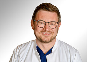 Porträt eines freundlich lächelnden Arztes mit Brille, Bart und kurz geschnittenen Haaren. Er trägt einen weißen Kittel über dunkelblauer Kleidung vor neutralem, hellem Hintergrund.