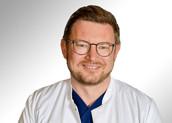 Porträt eines freundlich lächelnden Arztes mit Brille, Bart und kurz geschnittenen Haaren. Er trägt einen weißen Kittel über dunkelblauer Kleidung vor neutralem, hellem Hintergrund.