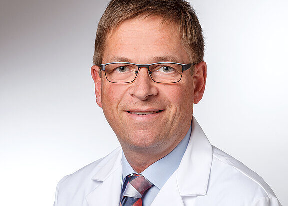 Kopf-und-Schulter-Porträt eines Mannes mit Brille im weißen Kittel, Hemd und Krawatte, freundlich lächelnd vor neutralem Hintergrund.