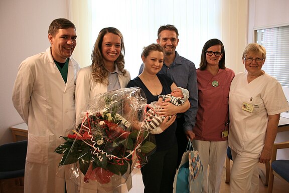 Eine Gruppe von sechs Personen steht in einem Krankenhauszimmer. Eine junge Frau hält ein neugeborenes Baby in den Armen, während eine andere Person einen Blumenstrauß präsentiert. Alle scheinen glücklich und stolz, die Atmosphäre ist feierlich und freundlich.