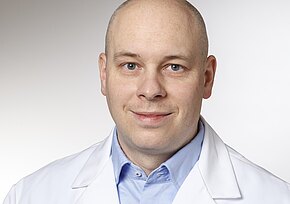 Ein mittelalter Mann mit Glatze trägt einen weißen Arztkittel über einem blauen Hemd. Er lächelt in die Kamera und hat blaue Augen. Der Hintergrund ist hell und neutral gehalten.