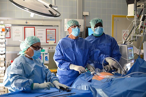 Operationsteam in blauen Kitteln, Hauben und Masken führt eine minimalinvasive Bauchoperation durch. Zwei Chirurgen arbeiten mit Laparoskopie-Instrumenten und beobachten den Monitor, eine Assistenz bereitet Instrumente vor. Steriler OP-Saal, Patient abgedeckt.