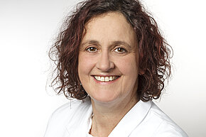 Susanne Dietze Porträt einer Frau mit lockigem, schulterlangem Haar in weißem Kittel, freundlich lächelnd, vor hellem, neutralem Hintergrund.