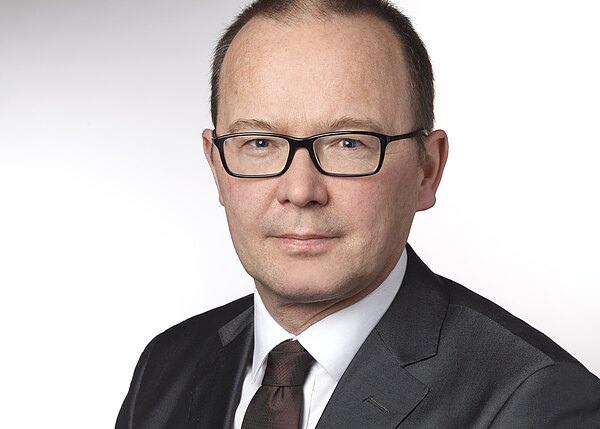 Professionelles Porträt: Mann mittleren Alters mit schwarzer Brille, kurzem Haar und neutralem Blick. Er trägt dunklen Anzug, weißes Hemd und dunkle Krawatte. Schulterporträt vor hellem, neutralem Hintergrund.